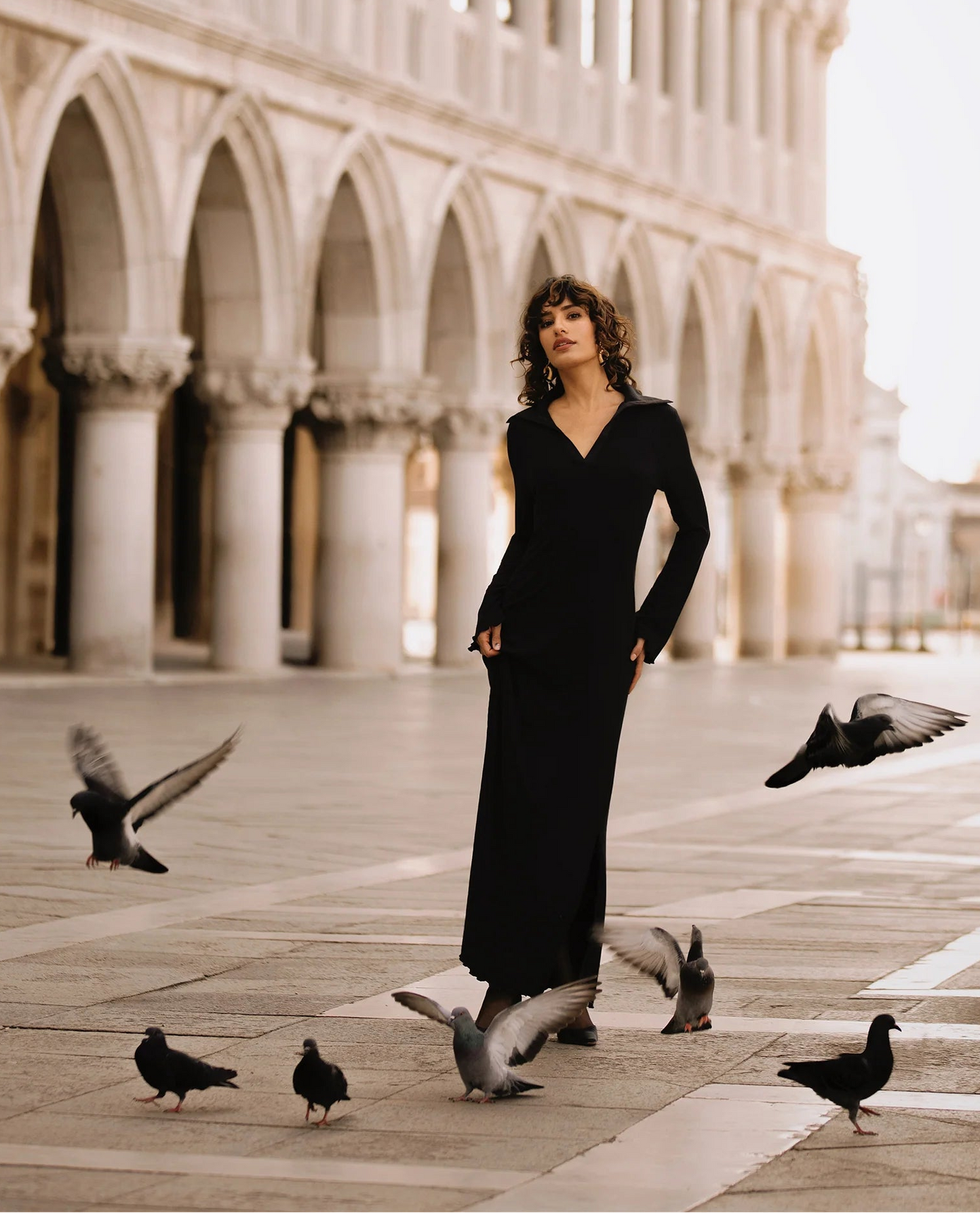 Woman in a black dress standing in front of an architectural structure with pigeons around her.