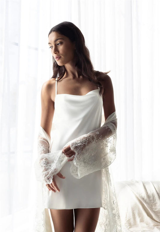 Woman wearing a white slip dress with lace sleeves against a white curtain background
