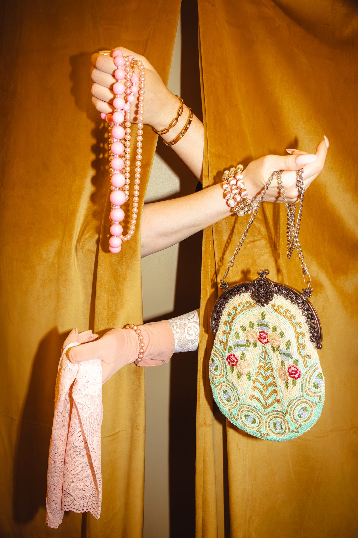 A person's hands and arms are shown holding fashion accessories in front of draped mustard-yellow fabric. The accessories include a light pink lace cloth, a strand of pink and beige pearls, a patterned green and cream embroidered clutch purse with a dark metal frame, and several gold-toned bracelets.