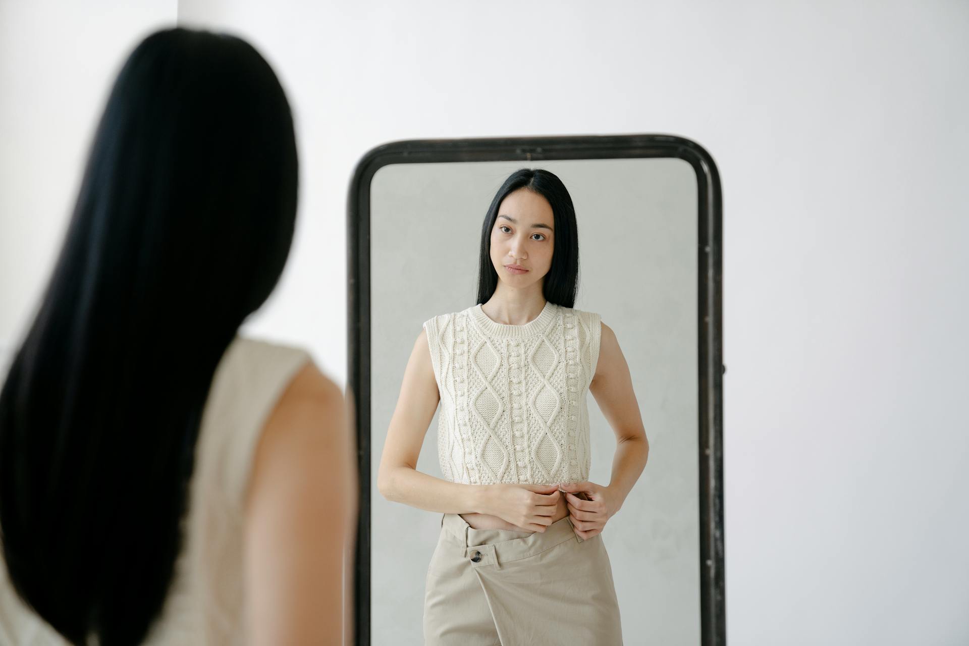 Person looking at themselves in a mirror wearing a white sleeveless top and beige pants.