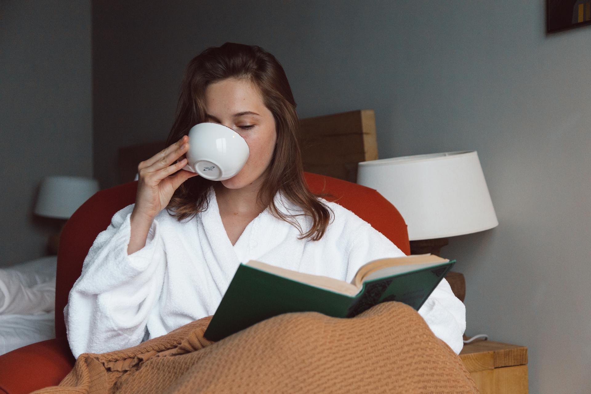 Woman in a robe drinking from a cup and reading a book in a room with lamps and a bed.