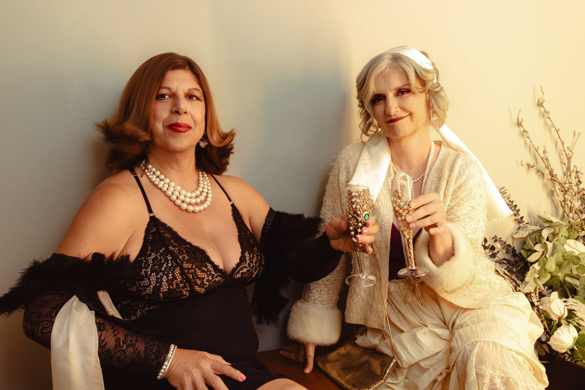 Two women posed together in vintage-inspired attire, one in a black lace dress with feathered sleeves and pearl necklace, the other in a cream 1920s-style flapper dress with fur trim and veil, each holding a champagne flute.