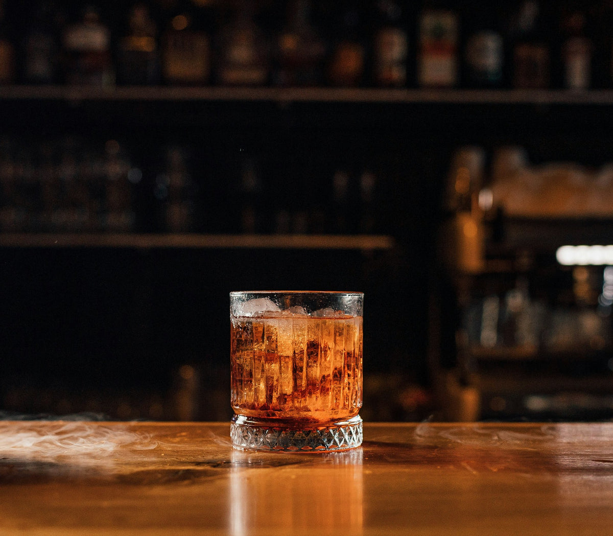 A clear rocks glass filled with an amber-brown cocktail and ice, set on a wooden bar counter with a blurred bar interior in the background.