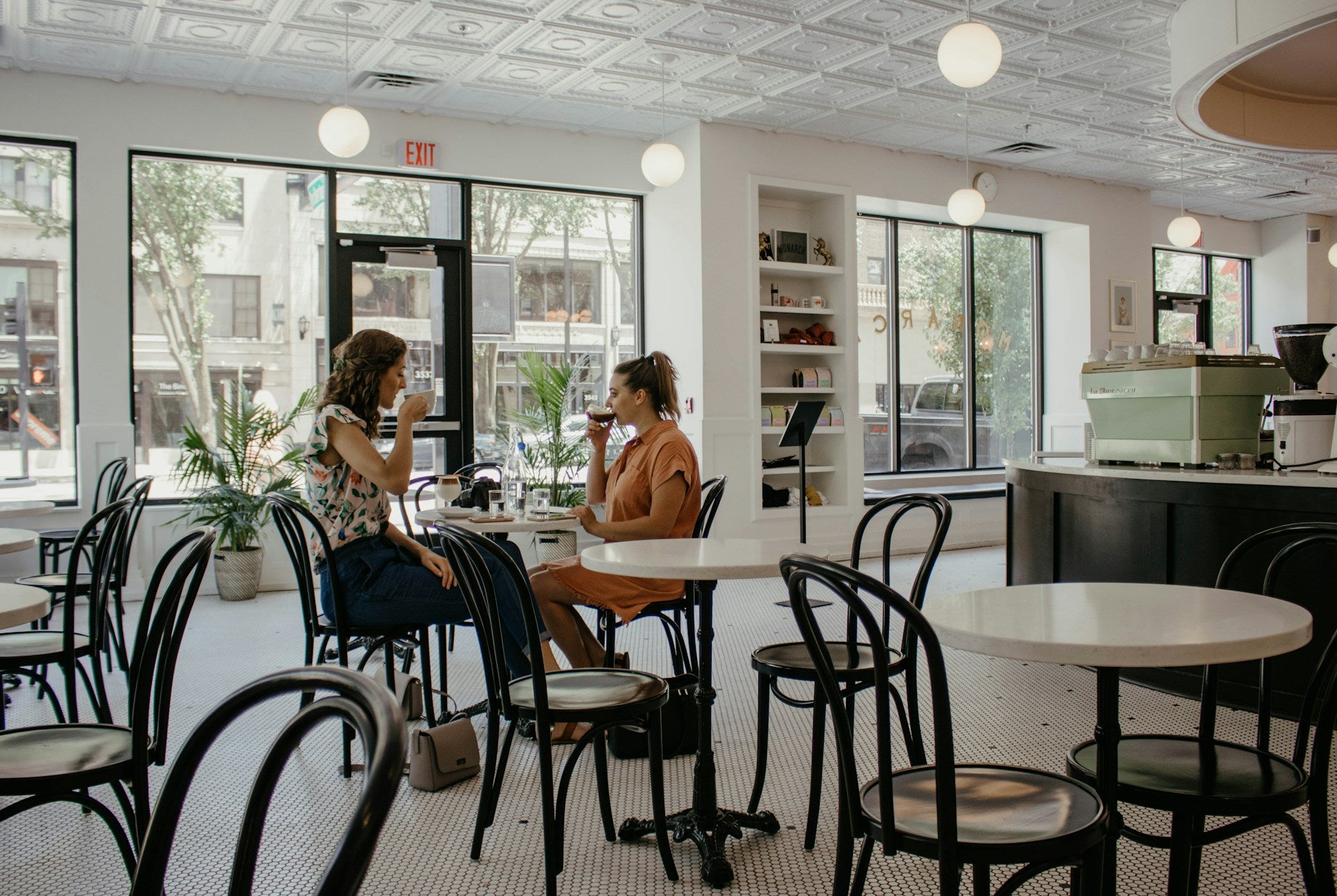 Two women sitting at a table in a modern cafe with large windows and a stylish interior.