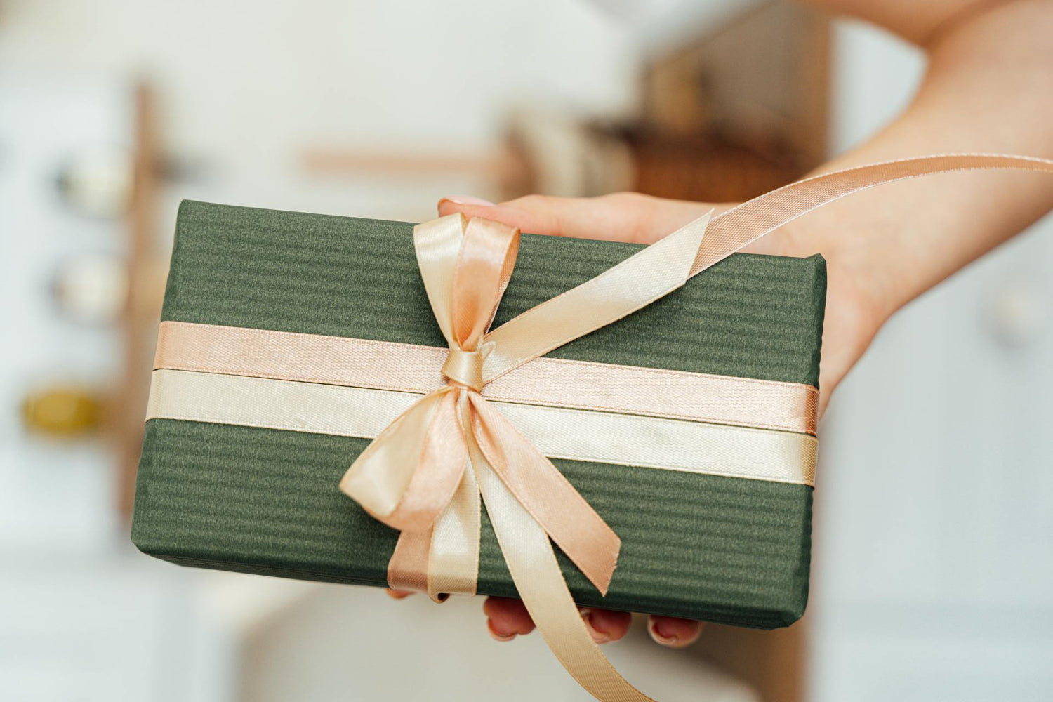 Green gift box with a pink ribbon held by a person against a blurred indoor background