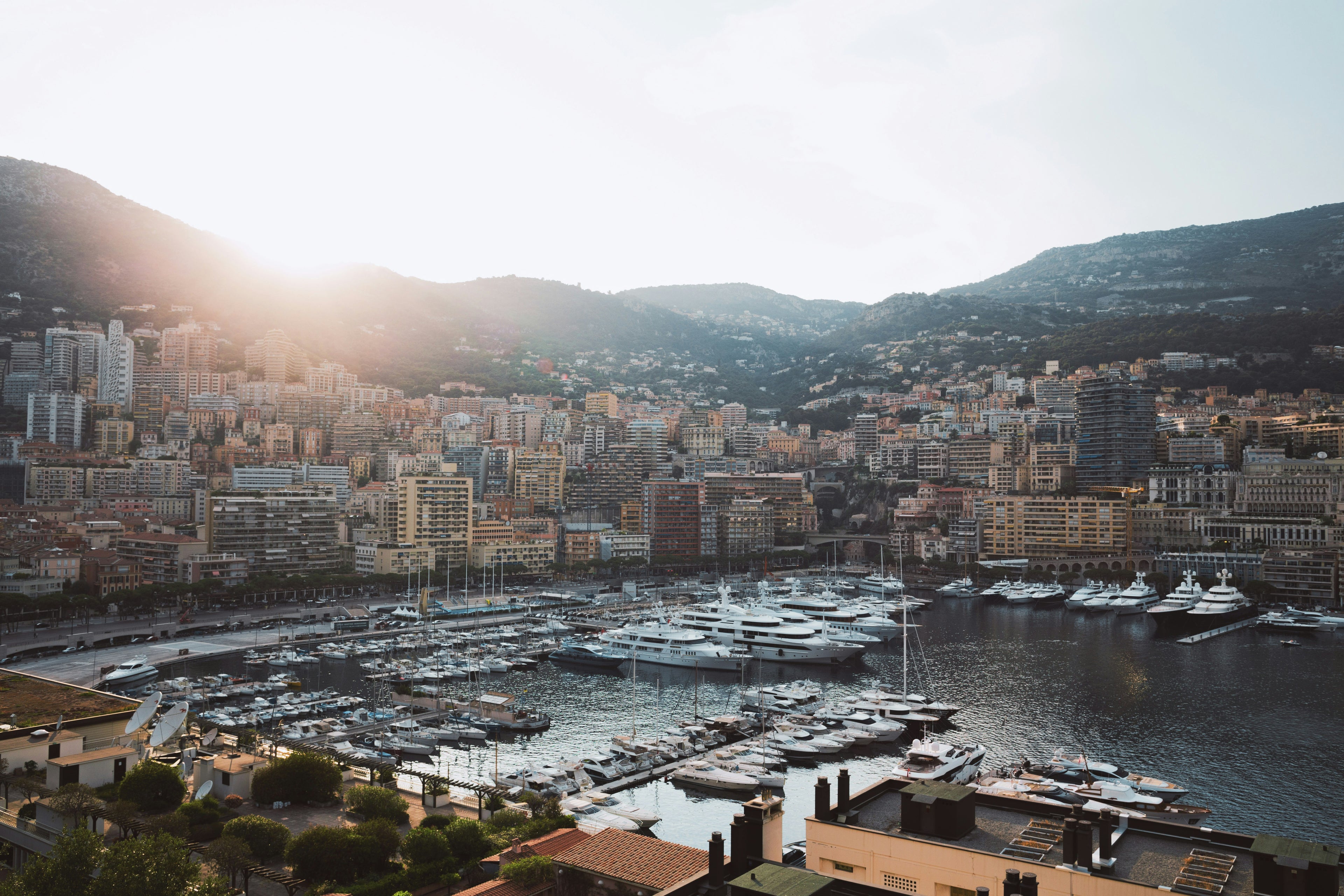Harbor scene with boats and buildings in a coastal city, sun setting over mountains.