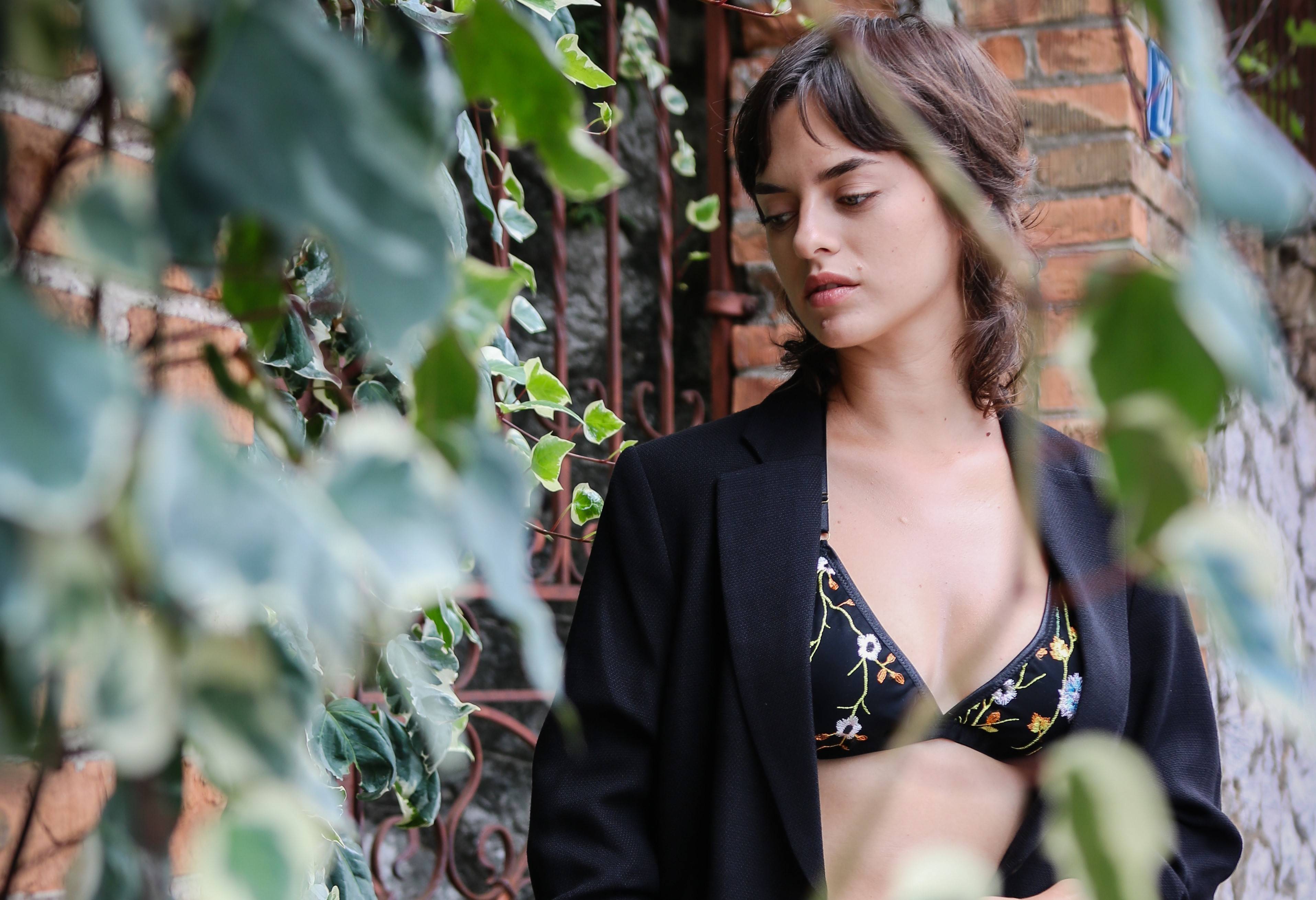 Woman wearing a black blazer over a floral top in an outdoor setting with greenery and a brick wall.
