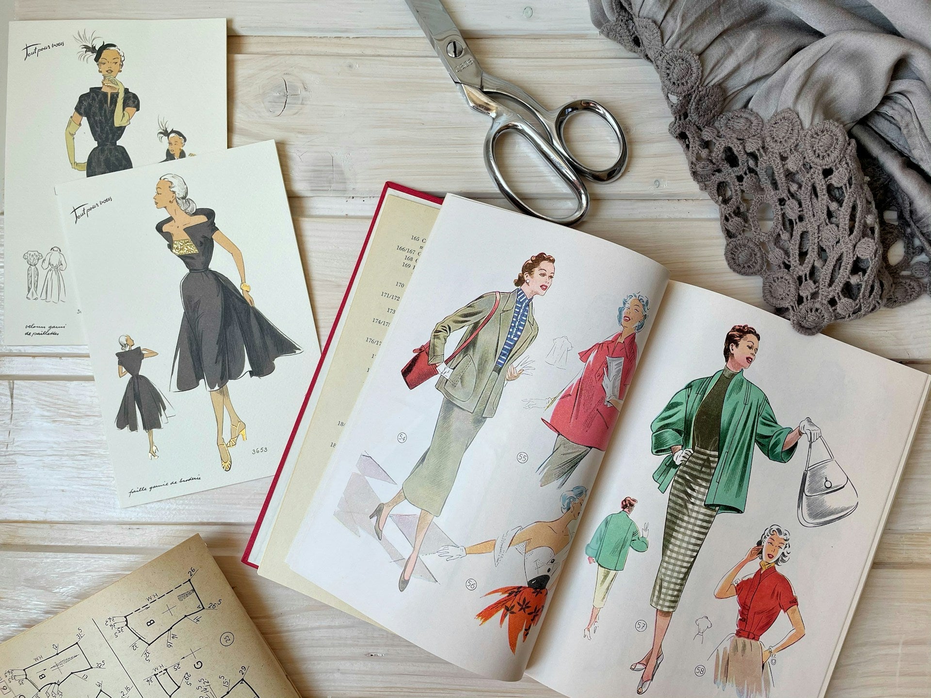 A styled flat-lay of vintage sewing patterns and fabric on a light wood surface, featuring an open book of illustrations, a card with dress sketches, silver scissors, and a gray lace-trimmed fabric.