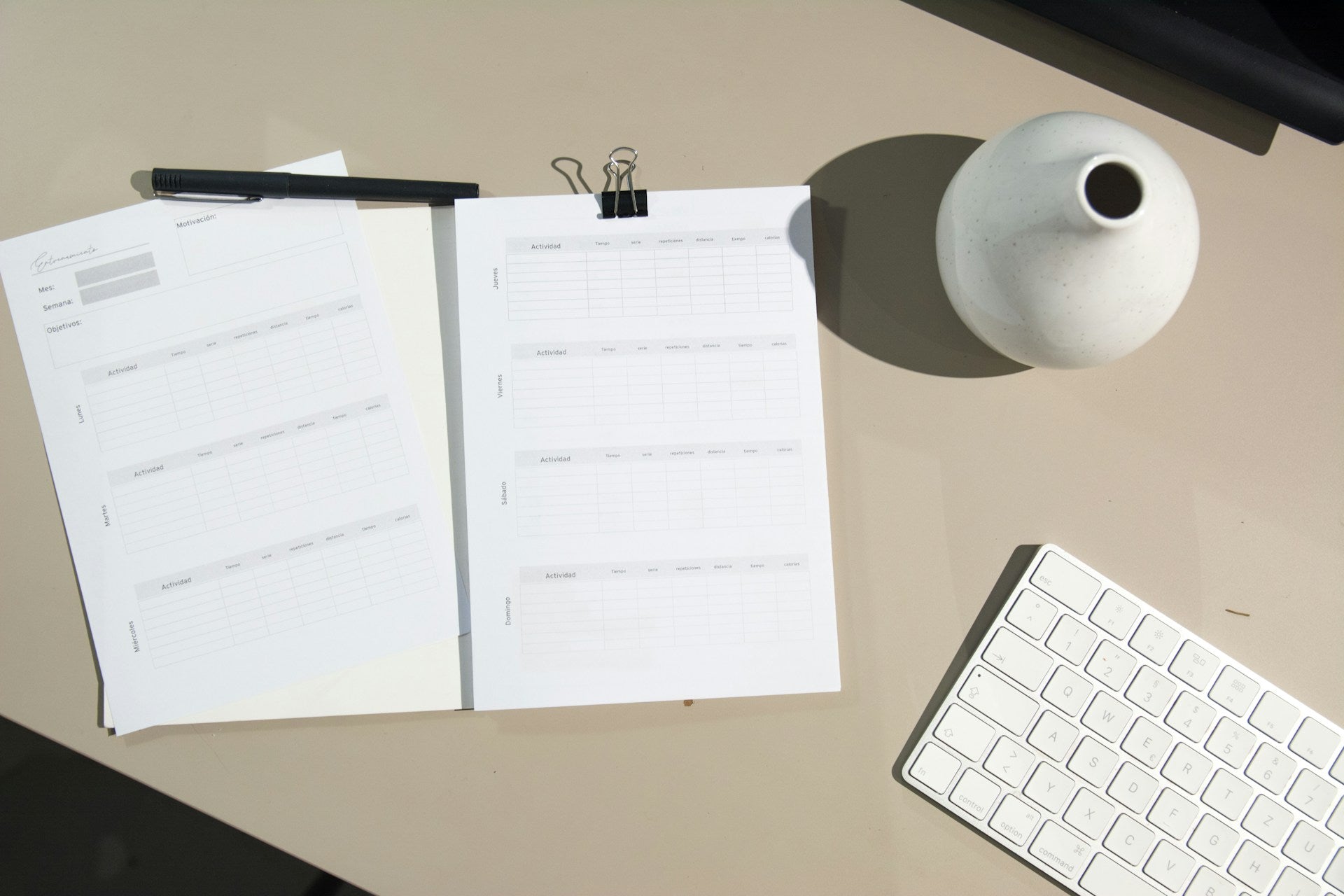 Open notebook with a pen on a desk next to a keyboard and a vase.